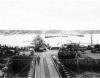Military vehicles (such as M3 Halftracks, an M8 Greyhound, and AFKWX Trucks) move ashore from Mulberry Artificial Harbor, across a pontoon bridge, to Omaha Beach.
