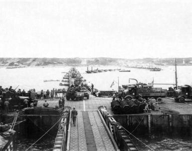 Military vehicles (such as M3 Halftracks, an M8 Greyhound, and AFKWX Trucks) move ashore from Mulberry Artificial Harbor, across a pontoon bridge, to Omaha Beach.