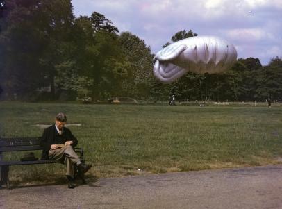 A man sits on a park bench in London, reading a book. Also a moored "barrage balloon" is visible in the background, while a second one soars high in the distance.